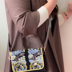 May include: A black crossbody bag with a flap closure. The bag features an embroidered floral design in shades of blue, yellow, and brown. The bag has a gold trim and a gray strap. The background is a brown coat.
