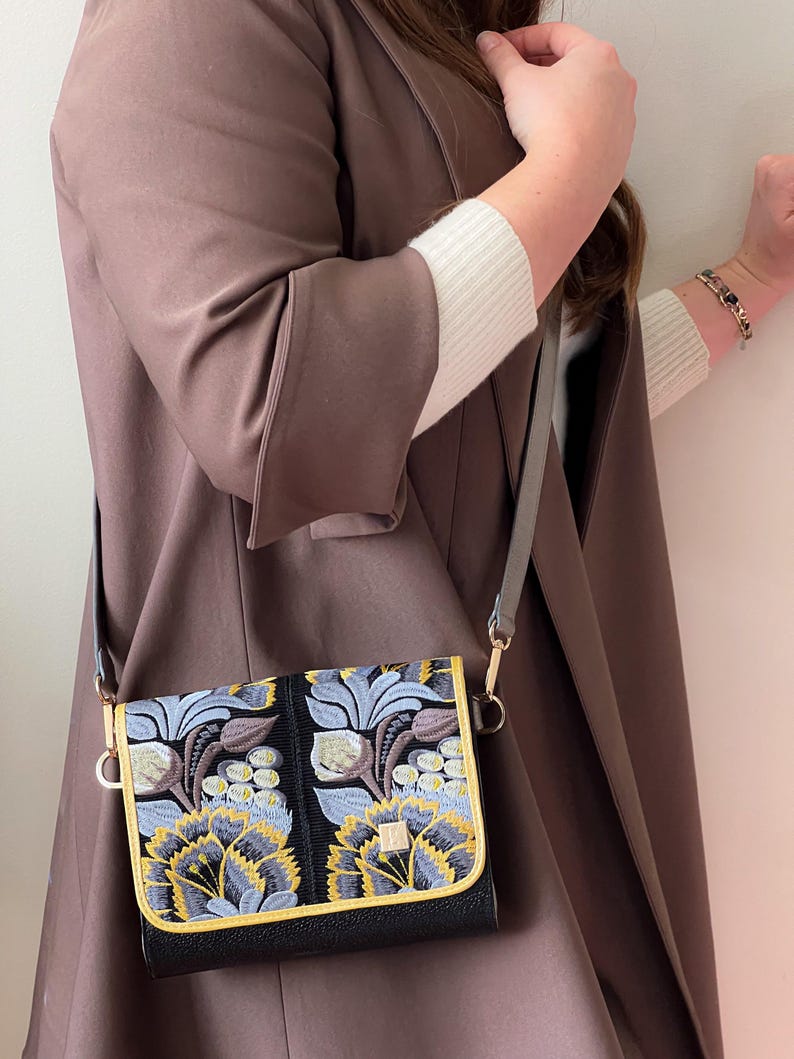 May include: A black crossbody bag with a flap closure. The bag features an embroidered floral design in shades of blue, yellow, and brown. The bag has a gold trim and a gray strap. The background is a brown coat.