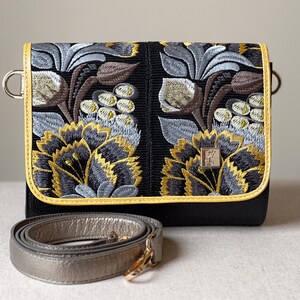 May include: Black crossbody bag with intricate floral embroidery in shades of gray, brown, and gold. The bag features a gold trim and a silver strap. The design includes a metal clasp and a decorative emblem.