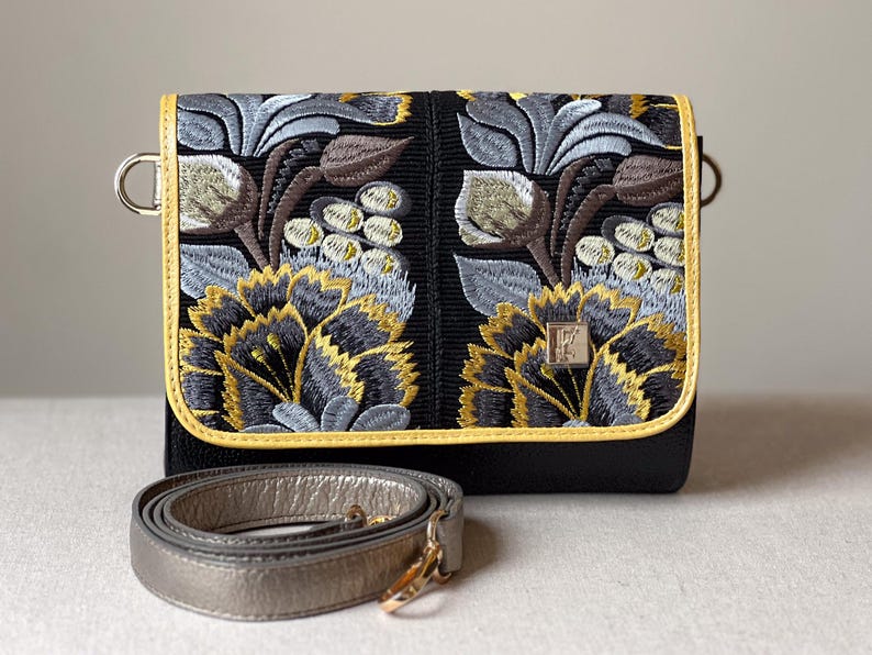 May include: Black crossbody bag with intricate floral embroidery in shades of gray, brown, and gold. The bag features a gold trim and a silver strap. The design includes a metal clasp and a decorative emblem.