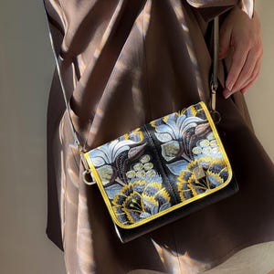 May include: Black crossbody bag with a floral design in yellow, blue, and white embroidery. The bag has a gold-colored trim and a thin strap. The bag is held by a person wearing a brown dress.