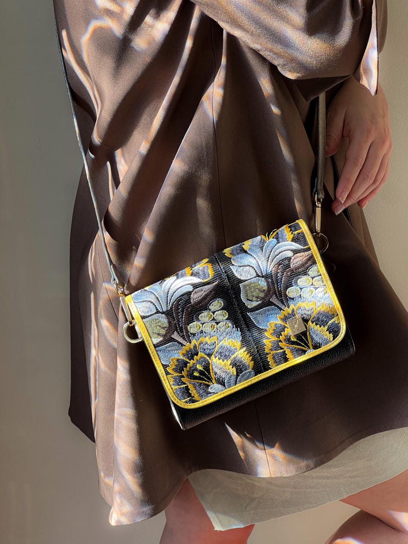 May include: Black crossbody bag with a floral design in yellow, blue, and white embroidery. The bag has a gold-colored trim and a thin strap. The bag is held by a person wearing a brown dress.