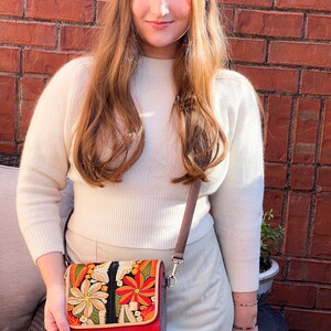 May include: A red crossbody bag with a colourful floral embroidered design. The bag features a brown strap and a rectangular shape. The embroidery includes orange, yellow, and green flowers. The bag is held by a person.