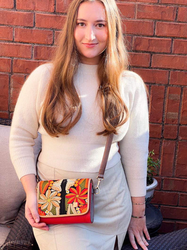 May include: A red crossbody bag with a colourful floral embroidered design. The bag features a brown strap and a rectangular shape. The embroidery includes orange, yellow, and green flowers. The bag is held by a person.