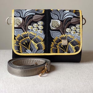 May include: Black crossbody bag with embroidered floral design in gold, gray, and brown. The bag has a gold-colored trim and a detachable silver strap. The bag's flap features a detailed floral pattern.