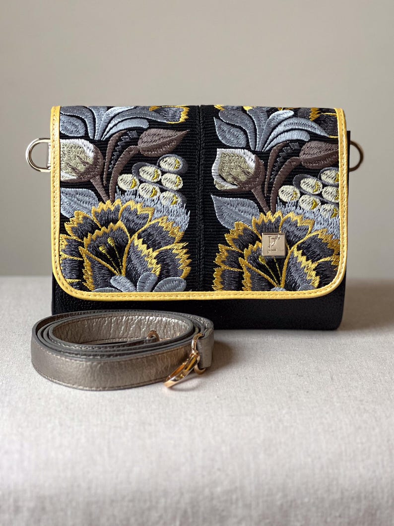 May include: Black crossbody bag with embroidered floral design in gold, gray, and brown. The bag has a gold-colored trim and a detachable silver strap. The bag's flap features a detailed floral pattern.