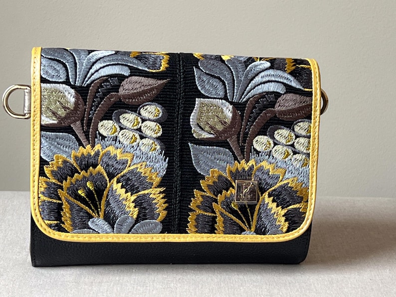 May include: Black clutch with embroidered floral design in yellow, gray, and brown. The bag has a yellow trim and a silver-colored metal clasp. The floral pattern is detailed and covers the front of the bag.