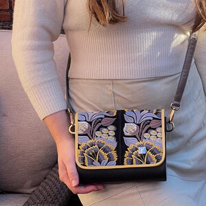 May include: Black crossbody bag with a flap closure, featuring an embroidered floral design in shades of purple, yellow, and gray. The bag has a gold trim and a brown strap. The bag is held by a person.