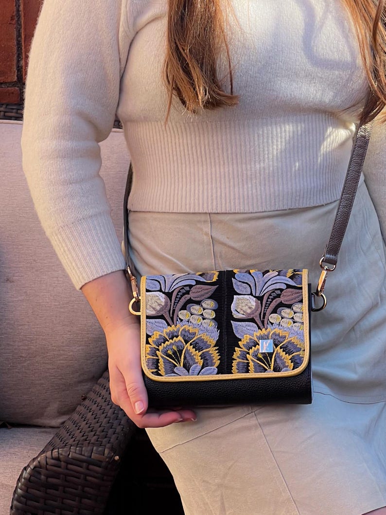 May include: Black crossbody bag with a flap closure, featuring an embroidered floral design in shades of purple, yellow, and gray. The bag has a gold trim and a brown strap. The bag is held by a person.