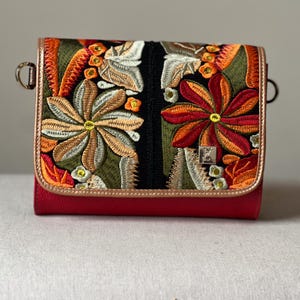 May include: A red crossbody bag with an embroidered floral design in orange, green, and cream. The bag features a flap closure, gold-coloured trim, and metal hardware. The bag's dimensions are not visible.