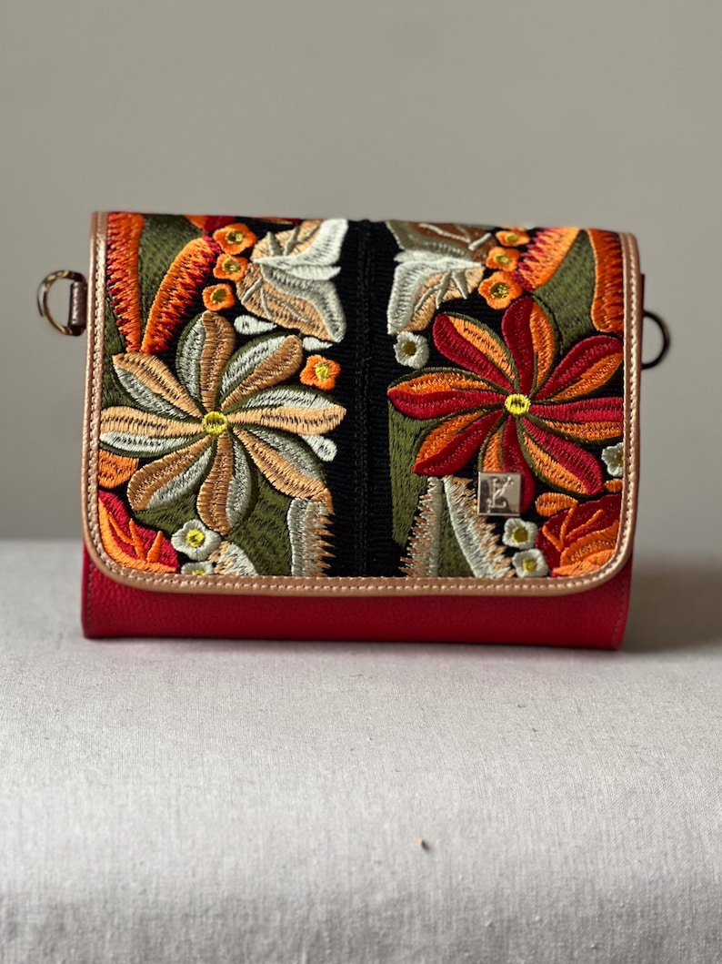 May include: A red crossbody bag with an embroidered floral design in orange, green, and cream. The bag features a flap closure, gold-coloured trim, and metal hardware. The bag's dimensions are not visible.
