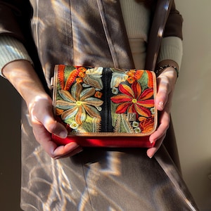 May include: A small, red handbag with a colourful floral embroidered design. The bag features a black background with orange, yellow, and green flowers. The bag is held in front of a brown coat.