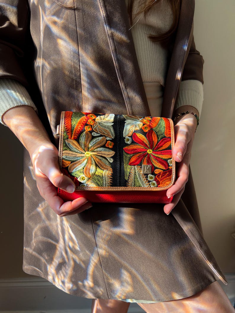 May include: A small, red handbag with a colourful floral embroidered design. The bag features a black background with orange, yellow, and green flowers. The bag is held in front of a brown coat.