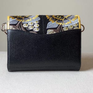 May include: Black crossbody bag with a flap featuring an embroidered floral design in gold, gray, and brown. The bag has a textured leather body and silver-tone hardware. The interior is not visible.