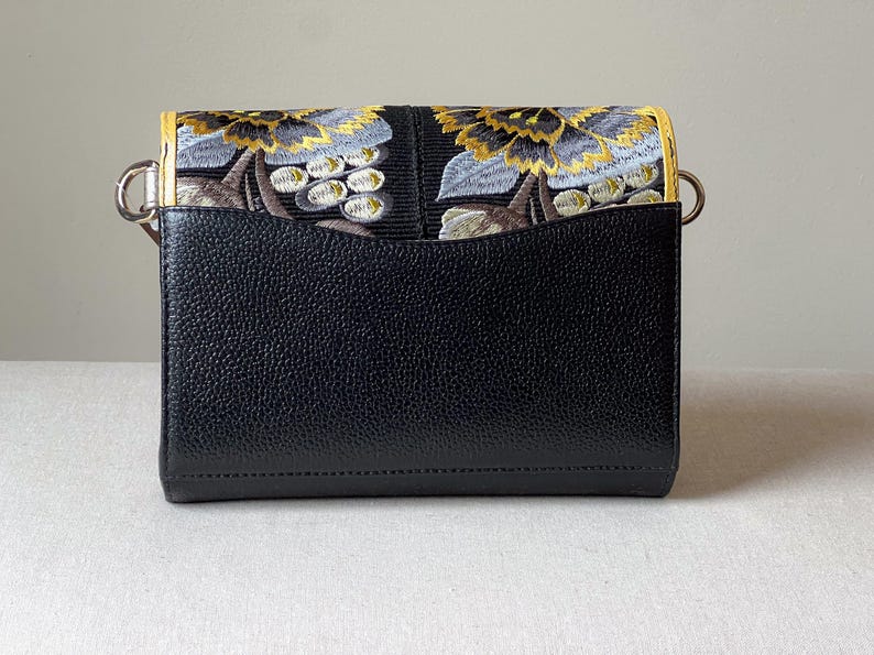 May include: Black crossbody bag with a flap featuring an embroidered floral design in gold, gray, and brown. The bag has a textured leather body and silver-tone hardware. The interior is not visible.