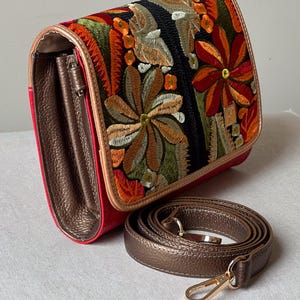 May include: A red crossbody bag with a floral embroidered flap. The flap has a black background with colourful embroidered flowers in shades of orange, green, and brown. A detachable brown strap and gold hardware complete the design.