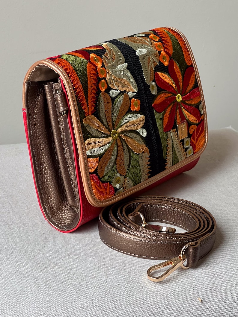 May include: A red crossbody bag with a floral embroidered flap. The flap has a black background with colourful embroidered flowers in shades of orange, green, and brown. A detachable brown strap and gold hardware complete the design.