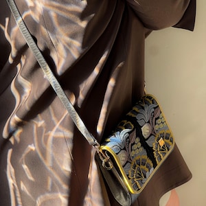 May include: A black crossbody bag with a floral pattern in blue, gold, and green. The bag has a gold trim and a gray strap. The bag is worn over a brown jacket.