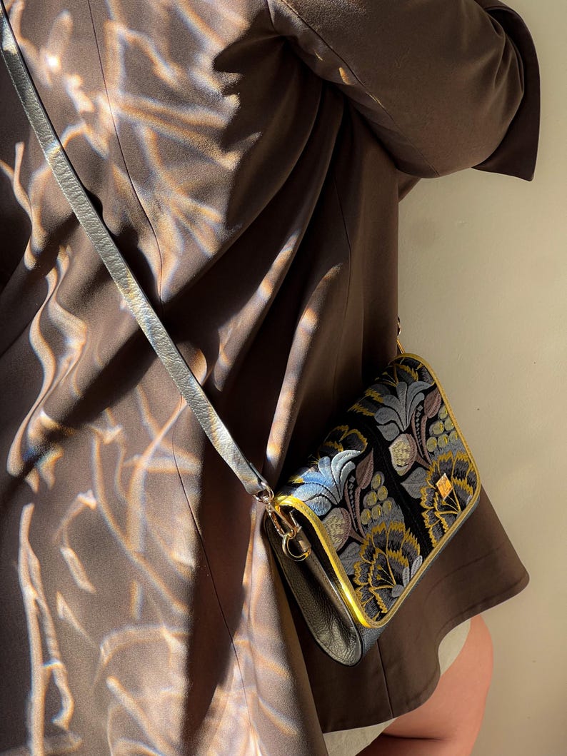 May include: A black crossbody bag with a floral pattern in blue, gold, and green. The bag has a gold trim and a gray strap. The bag is worn over a brown jacket.