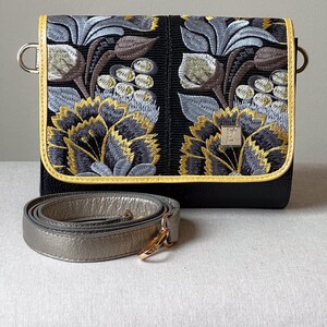 May include: Black crossbody bag with floral embroidery in shades of yellow, gray, and brown. The bag has a yellow trim and a silver strap. The bag features a silver clasp and metal hardware.