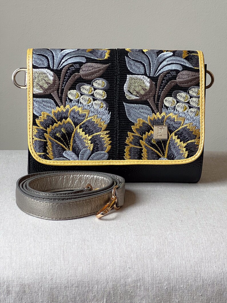 May include: Black crossbody bag with floral embroidery in shades of yellow, gray, and brown. The bag has a yellow trim and a silver strap. The bag features a silver clasp and metal hardware.