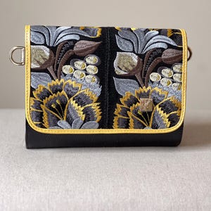 May include: Black crossbody bag with embroidered floral design in yellow, gray, and brown. The bag features a yellow trim and a silver metal clasp. The floral pattern is centered on the front of the bag.