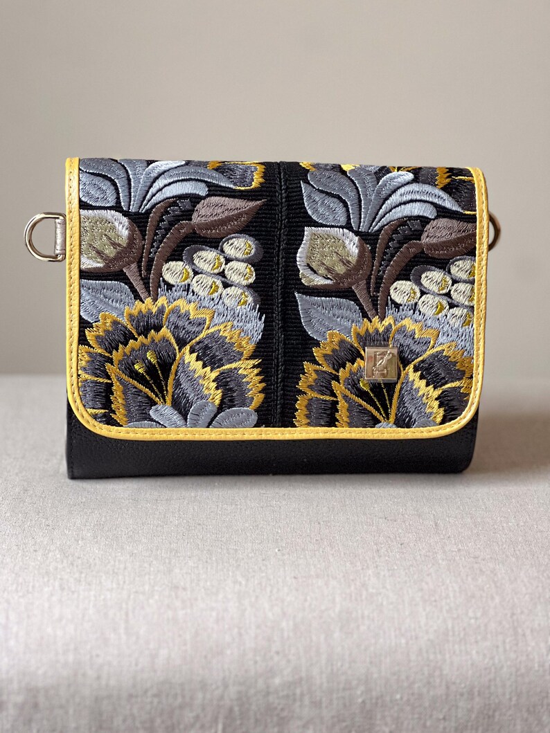 May include: Black crossbody bag with embroidered floral design in yellow, gray, and brown. The bag features a yellow trim and a silver metal clasp. The floral pattern is centered on the front of the bag.