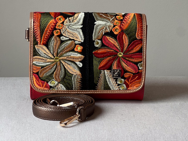 May include: A red handbag with a floral embroidered design in shades of orange, green, and brown. The bag has a brown leather strap and gold-coloured hardware. The flap closure is outlined in brown leather.