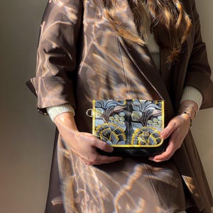 May include: A small, black crossbody bag with an embroidered floral design in shades of blue, yellow, and gray. The bag has a gold trim and a black strap. The person is wearing a brown blazer.