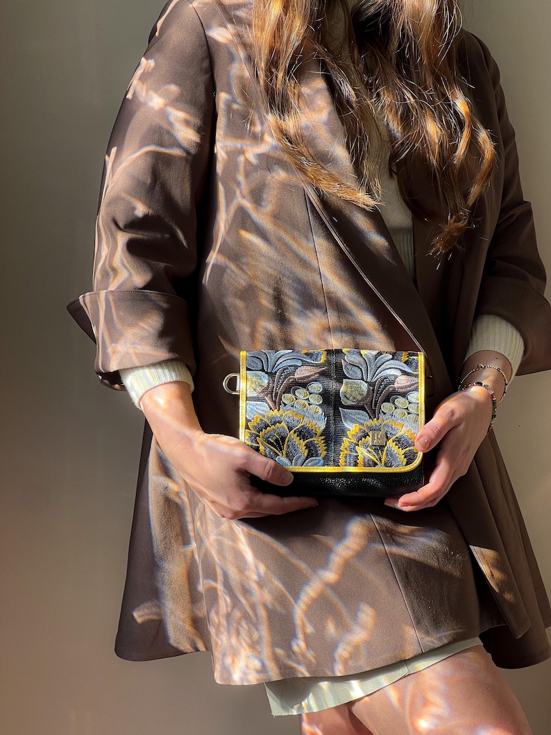 May include: A small, black crossbody bag with an embroidered floral design in shades of blue, yellow, and gray. The bag has a gold trim and a black strap. The person is wearing a brown blazer.