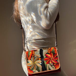 May include: A rectangular crossbody bag with a brown leather strap and trim. The bag features a vibrant floral embroidery design in shades of orange, red, green, and yellow against a black background. The bag is approximately 23 cm wide.