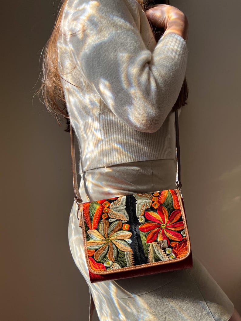 May include: A rectangular crossbody bag with a brown leather strap and trim. The bag features a vibrant floral embroidery design in shades of orange, red, green, and yellow against a black background. The bag is approximately 23 cm wide.