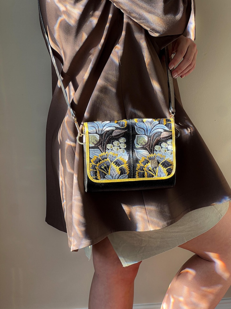 May include: A black crossbody bag with a floral pattern in blue, yellow, and white. The bag has a gold trim and a silver strap. The bag is rectangular and appears to be made of a textured material.
