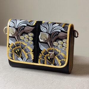 May include: Black crossbody bag with embroidered floral design in gray, yellow, and brown. The bag features a gold trim and a silver clasp. The floral pattern is detailed and covers the front of the bag.