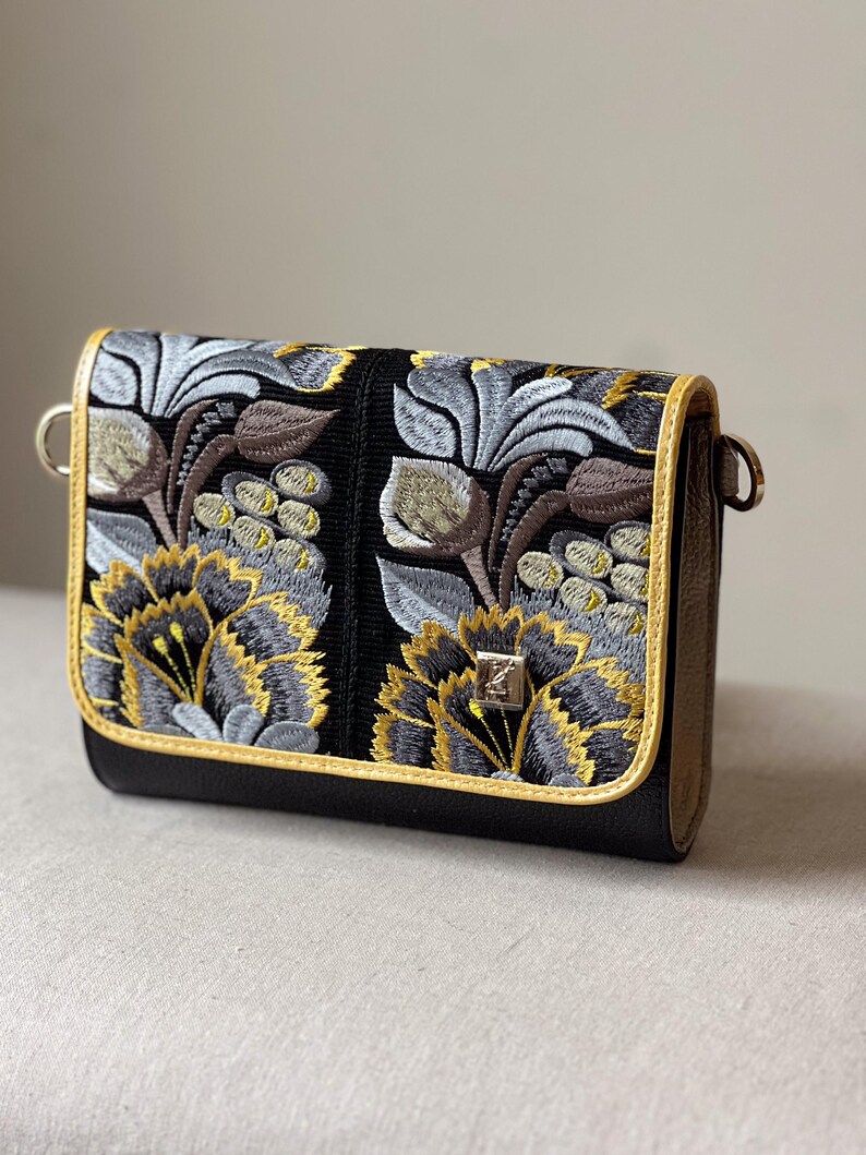May include: Black crossbody bag with embroidered floral design in gray, yellow, and brown. The bag features a gold trim and a silver clasp. The floral pattern is detailed and covers the front of the bag.