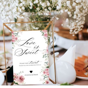 May include: A white sign with pink floral borders and the text "Love is Sweet" with a heart and the names "Ayanna & Omar". The sign is in a gold frame and is on a table with white flowers.
