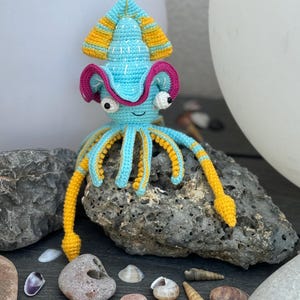 May include: A handmade crochet squid toy in shades of blue, yellow, and pink. The squid has large, expressive eyes and a smiling mouth. The tentacles are yellow and blue. The toy is displayed on a rock with seashells.