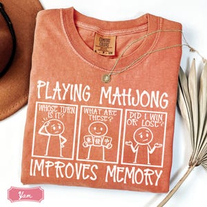 May include: A terracotta-colored t-shirt featuring a white graphic with the text "PLAYING MAHJONG IMPROVES MEMORY" and three panels showing stick figures asking "WHOSE TURN IS IT?", "WHAT ARE THESE?", and "DID I WIN OR LOSE?" with mahjong tiles.