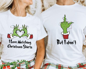 Può includere: T-shirt bianche con grafiche a tema natalizio. Una maglietta presenta mani verdi del Grinch che formano un cuore con la scritta "I Love Matching Christmas Shirts". L'altra maglietta ha una mano del Grinch che fa il dito medio con la scritta "But I don't."
