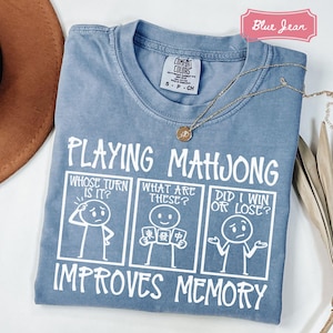 May include: A blue jean colored t-shirt featuring a white graphic of three comic panels with stick figures playing mahjong. The text reads Playing Mahjong Improves Memory, with panels asking Whose Turn Is It, What Are These, and Did I Win Or Lose.
