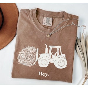 May include: A brown t-shirt with a white graphic of a tractor pulling a bale of hay. The text "Hey." is printed below the graphic.