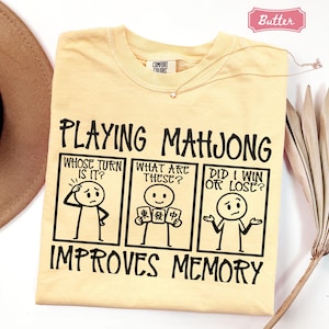May include: A butter yellow t-shirt featuring a black graphic of a three-panel comic strip with stick figures. The text reads: PLAYING MAHJONG, WHOSE TURN IS IT?, WHAT ARE THESE?, DID I WIN OR LOSE?, and IMPROVES MEMORY. The shirt has a Comfort Colors label.