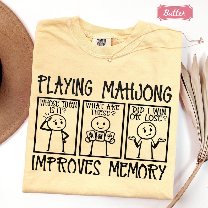 May include: A butter yellow t-shirt featuring a black graphic of a three-panel comic strip with stick figures. The text reads: PLAYING MAHJONG, WHOSE TURN IS IT?, WHAT ARE THESE?, DID I WIN OR LOSE?, and IMPROVES MEMORY. The shirt has a Comfort Colors label.