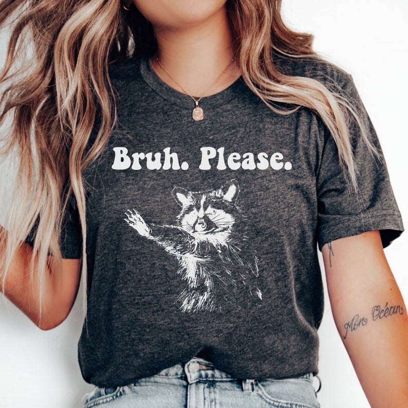 May include: A dark grey t-shirt with a white graphic of a raccoon with the text "Bruh. Please." printed on the front.