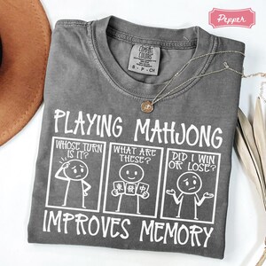 May include: A charcoal gray t-shirt featuring white graphic text that reads Playing Mahjong Improves Memory. The design includes three comic-style panels with stick figures asking Whose Turn Is It, What Are These, and Did I Win Or Lose.