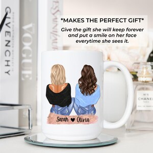 May include: White ceramic mug featuring illustrations of two women with blonde and brown hair, wearing black and blue tops and jeans. The mug has the names "Sarah" and "Olivia" printed on it. The mug is on a mirrored coaster.