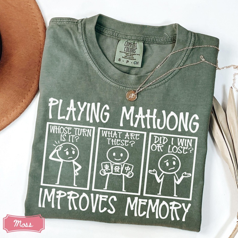 May include: A moss green short-sleeve t-shirt featuring a white graphic with three panels depicting stick figures and the text Playing Mahjong Improves Memory. The panels include the questions Whose Turn Is It, What Are These, and Did I Win Or Lose.