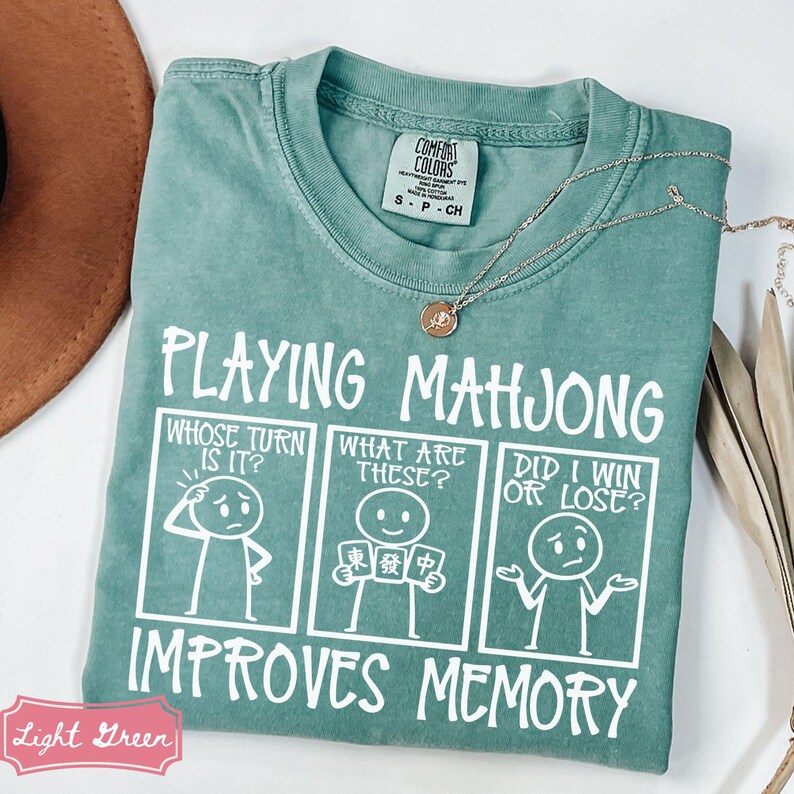 May include: A sage green t-shirt featuring a white graphic print with the text "PLAYING MAHJONG IMPROVES MEMORY" and three panels showing stick figures asking "WHOSE TURN IS IT?", "WHAT ARE THESE?", and "DID I WIN OR LOSE?" with mahjong tiles.