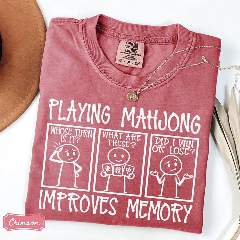 May include: A crimson short-sleeved t-shirt featuring white graphic text and three stick figure panels. The text reads Playing Mahjong Improves Memory, with panels asking Whose Turn Is It, What Are These, and Did I Win Or Lose. A gold necklace rests on the collar.
