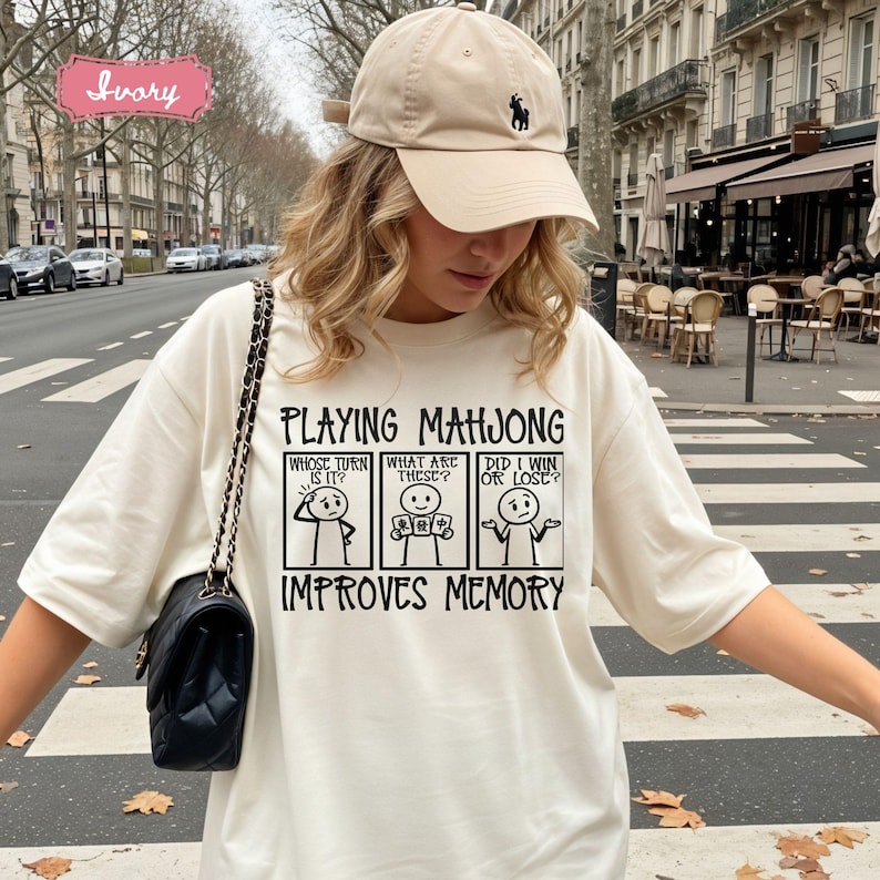 May include: An ivory t-shirt featuring a black graphic print of three stick figure panels with the text Playing Mahjong Improves Memory. The panels include the questions Whose turn is it, What are these, and Did I win or lose. The shirt is paired with an ivory baseball cap.